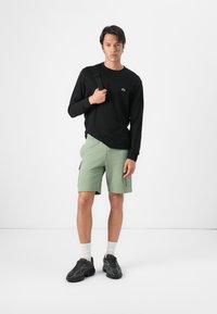 Schwarzes Langarmshirt mit grünem Logo, kombiniert mit hellgrünen Shorts und schwarzen Sportschuhen. Modell hält einen Riemen und steht vor einem weißen Hintergrund.