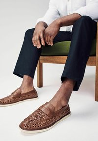 Zapatos marrones de cuero tipo slip-on con diseño trenzado, con suela plana y plantilla acolchada. Usados con pantalones negros, sentados en una silla de madera.