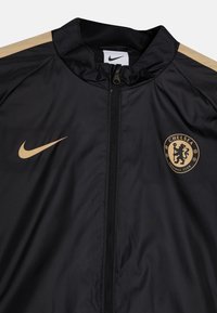 Chaqueta del Chelsea Football Club de Nike en negro con acentos dorados. Presenta un cuello alto, cierre frontal y el logo del club en el pecho.
