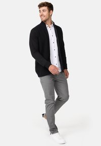 Cardigan noir en tricot avec un col, superposé sur une chemise blanche à boutons, associé à un pantalon gris clair et des baskets blanches.