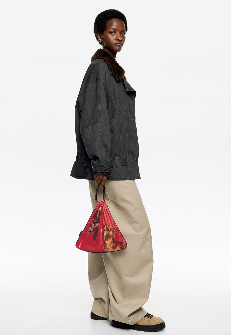 Przesadzona ciemnoszara kurtka z brązowym futrzanym kołnierzem, beżowe szerokie spodnie i czerwona torba na sznurkach z nadrukiem psów. Brązowe i czarne obuwie.