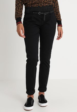 Zwarte katoenen broek met een slanke pasvorm, voorzien van een riem en opgerolde boorden, gecombineerd met zwarte sneakers en een bruine trui met patroon.