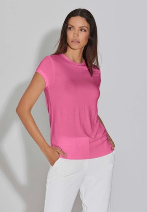 Frau mit langen braunen Haaren, die ein eng anliegendes pinkfarbenes kurzärmliges Hemd und weiße Hosen trägt, steht mit den Händen in den Taschen und schaut zur Seite.