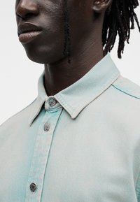 Ανοιχτό μπλε πουκάμισο από τζιν με μυτερό γιακά, διακοσμημένο με ασημί κουμπιά και ελαφρώς υφασμένο ύφασμα. Κοντινή άποψη του λαιμού.