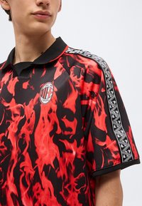 Νεαρό άτομο που φοράει μαύρη και κόκκινη ποδοσφαιρική φανέλα της AC Milan με φλόγες και ρίγες με ασπρόμαυρο σχέδιο στον ώμο.