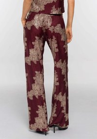 Mujer vestida con pantalones de pierna ancha burdeos con patrones florales metálicos dorados y zapatos de tacón negros, de pie con las manos en los bolsillos.