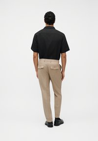 Homme portant un pantalon beige sur mesure avec des poches arrière boutonnées, une chemise noire à manches courtes et des chaussures noires, debout devant un fond blanc uni.