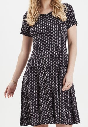 Mujer con vestido negro hasta la rodilla, de mangas cortas y pequeño estampado de hojas rosas, de pie frente a un fondo blanco.