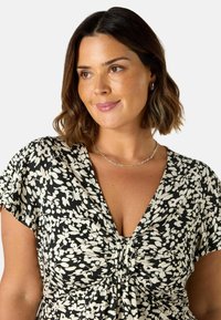 Czarna i biała sukienka w kwiaty z głębokim dekoltem w kształcie V i krótkimi rękawami, z detalem w postaci marszczonego materiału na środku. Srebrny naszyjnik łańcuszkowy.