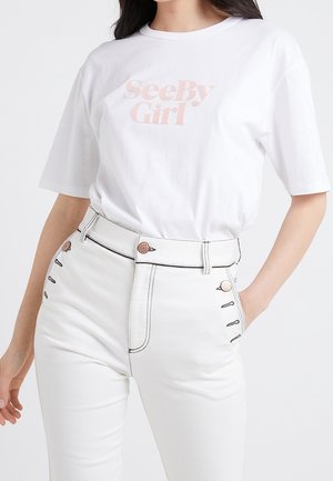 Kobieta w białym t-shircie z krótkim rękawem i jasnoróżowym napisem „See By Girl”, włożonym w białe spodnie z wysokim stanem i czarnymi przeszyciami.