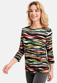 Lächelnde Frau, die ein bunt gestreiftes Langarmshirt und graue Hosen trägt, geht mit ihrem linken Arm leicht ausgestreckt.