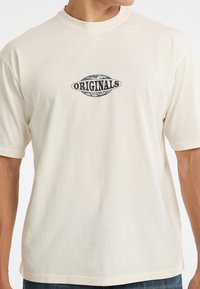 T-shirt a maniche corte color crema in cotone, con un logo grafico centrale in nero e blu che legge "ORIGINALS GUESS."
