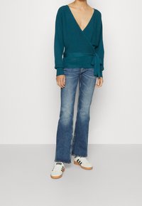 Teal wikkeltrui met diepe V-hals en strik in de taille, gecombineerd met uitlopende blauwe jeans en witte sneakers met zwarte accenten.