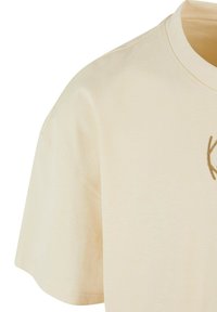 T-shirt en coton beige clair avec un col rond, des manches courtes et un petit logo doré brodé sur la poitrine. Texture douce.