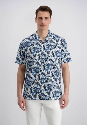 Man draagt een kortemouw, knoopjes overhemd met blauw-wit bloemenpatroon en witte broek, staand tegen een effen achtergrond.