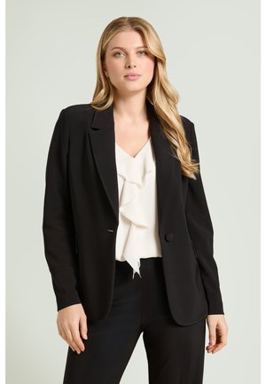 Femme aux longs cheveux blonds portant un blazer noir et un pantalon avec un chemisier blanc à volants, debout devant un fond clair uni.