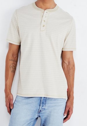 Mann, der ein beigefarbenes, kurzärmliges Henley-Shirt mit dezenten Streifen und hellblauen Jeans trägt, mit Tattoos an beiden Unterarmen.