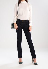 Blusa blanca con puños con volantes, jeans ajustados de color azul oscuro con botones dorados, bolso verde y tacones verdes contra un fondo neutro.