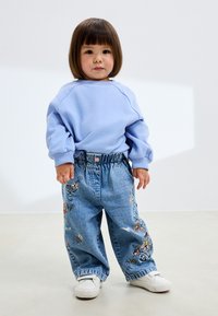 Világoskék pulóver bordázott mandzsettákkal, laza, hímzett farmerral, amely virágmintákat visel, fehér tornacipővel párosítva.