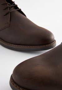 Hnedé kožené topánky s hladkou textúrou, zaoblenou špičkou a minimalistickým dizajnom. Sú viditeľné šnúrky a detaily šitia.