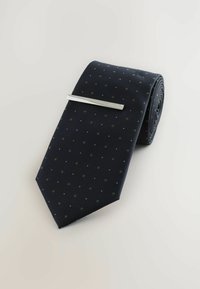 Maritimer Krawatte mit kleinen grünen und weißen Punkten, aus strukturiertem Stoff, ordentlich gerollt mit einer silbernen Krawattennadel, die quer darüber positioniert ist.
