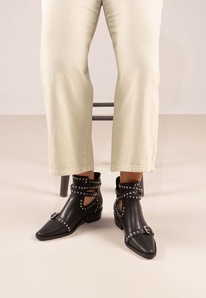 Botas negras hasta el tobillo con puntera afilada, correas con tachuelas y un tacón bajo. Confeccionadas en cuero suave con detalles recortados.