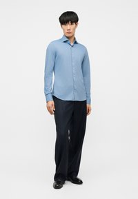 Νεαρός άνδρας που στέκεται, φορώντας γαλάζιο πουκάμισο με κουμπιά, σκούρο παντελόνι με ίσιες ρίγες και μαύρα παπούτσια επίσημου τύπου, μπροστά σε λευκό φόντο.