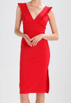Kobieta w czerwonej sukience bez rękawów z falbankami na ramiączkach, głębokim dekoltem w kształcie litery V, dopasowaną talią i rozcięciem po boku, stojąca na jednolitym tle.