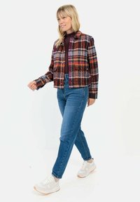 Kariertes, kürzes Jacket in vielseitigen Farben, einschließlich Rot, Braun und Blau, kombiniert mit blauen Jeans und hellen Sneakers. Schlichter Hintergrund.