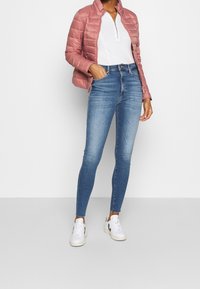 Ljusrosa kviltad puffjacka, vit skjorta med krage, blå smala jeans och vita sneakers med svarta detaljer. Enkla bakgrund.