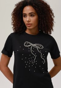 Sort T-shirt med perleudsmykninger i en bue-design, med spredte perler der strækker sig nedad fra buen. Korte ærmer, rund halsudskæring.