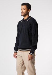 Polo a maniche lunghe nera in maglia con texture a coste, caratterizzata da colletto e polsini a righe, abbinata a pantaloni beige chiaro.