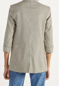 Geruite blazer in beige en zwart, met driekwart mouwen en een achterventilatie. Het materiaal lijkt gestructureerd en is ontworpen met een geruit patroon.