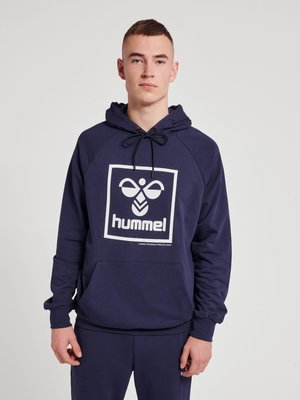 Jonge man in een marineblauwe hummel hoodie met groot logo en bijpassende marineblauwe broek, staand tegen een effen lichte achtergrond.
