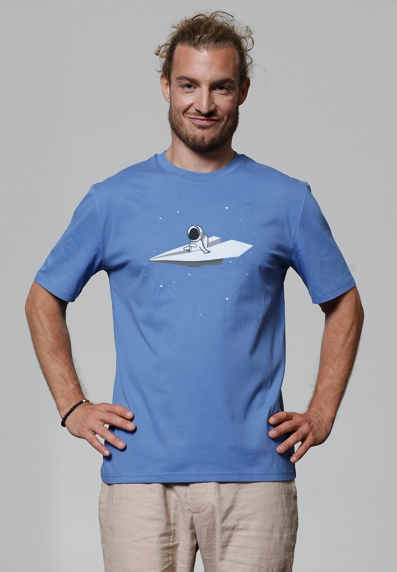 T-shirt en coton bleu avec un motif d'astronaute sur un avion en papier, sur fond étoilé simple. Manches courtes, col rond.