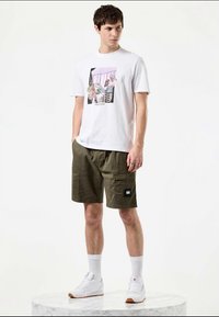 T-shirt gráfica branca com uma estampa colorida, combinada com calções cargo verde-oliva com dois bolsos laterais e um rótulo preto. Sneakers brancos.