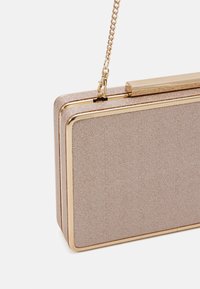 Beige rektangulär kuvertväska med texturerad yta, guldram och avtagbar guldkedjestrar som är fäst i det övre hörnet.