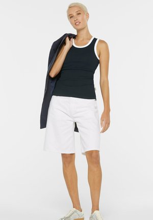 Frau mit kurzen blonden Haaren, die ein schwarzes Tanktop mit weißem Rand, weiße knielange Shorts und silberne Sneaker trägt, hält eine dunkle Jacke über der Schulter.