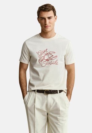 Young man wearing cream T-shirt with red lobster graphic and "Gant Seaside" text, paired with cream pants and dark braided belt.