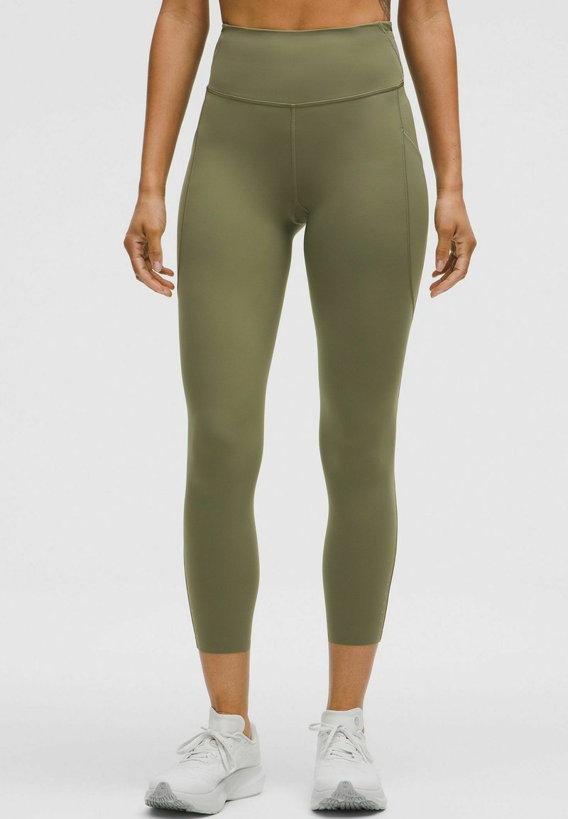 Olivgrüne, hochgeschnittene Leggings aus einem glatten, dehnbaren Stoff; enganliegendes Design und dezente Seitennähte; kombiniert mit weißen Sportschuhen.