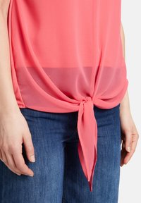 Blusa corallo realizzata in tessuto leggero e trasparente, con un nodo annodato nella parte inferiore e un design a strati, abbinata a jeans in denim blu.