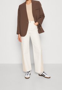 Blazer marrón sobre top beige, pantalones blancos de pierna ancha y zapatillas blancas con rayas negras. Detalle notable de bolsillo en el blazer.