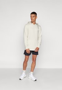 Gai smilšu krāsas kapuce ar priekšējo kabatu un aukliņu, apvienota ar tumšiem īsiem šortiem un baltām sporta apavām. Audums izskatās gluds.