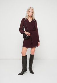 Μπορντό περιτύλιγμα φόρεμα με μακριά μανίκια, V-λαιμόκοψη και ζωνάκι, συνδυασμένο με μαύρες δερμάτινες μπότες στιλέτο μέχρι το γόνατο και μια μικρή τσάντα ώμου.