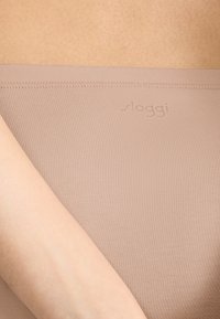 Beige nahtlose Stretch-Slips mit gerippter Textur. Der Bund zeigt ein dezentes "sloggi"-Logo. Das Bild zeigt eine teilweise Ansicht mit einem Arm.
