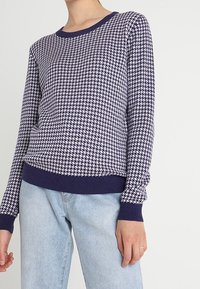 Námorně modrý pletený svetr s geometrickým vzorem, kulatým výstřihem a náplety; spuštěný s světle modrými rovnými džíny.