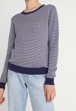 Granatowy sweter dzianinowy z geometrycznym wzorem, okrągłym dekoltem i ściągaczami; w połączeniu z jasnoniebieskimi dżinsami o prostym kroju.