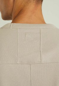 Beige crew neck t-shirt features ribbed texture, visible stitching, and an embroidered triangular detail at the back neckline.