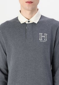 Siva polo majica z dolgimi rokavi in kremnim ovratnikom, ki vsebuje zaponko na gumb in vezeno "H" logo na levem prsnem delu.