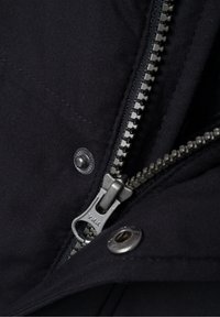 Tissu de veste noire avec une fermeture éclair métallique YKK visible, dotée d'un bouton-pression et de coutures détaillées. Texture douce et design durable.